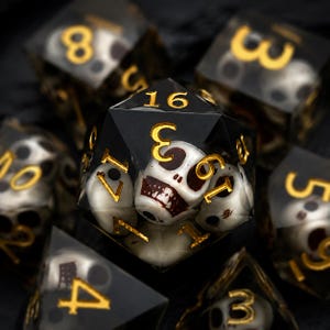 May include: A set of polyhedral dice made of black resin with embedded white skull figures inside each piece. The numbers are engraved in a metallic gold font. The central twenty-sided die prominently displays the number 16 at the top.