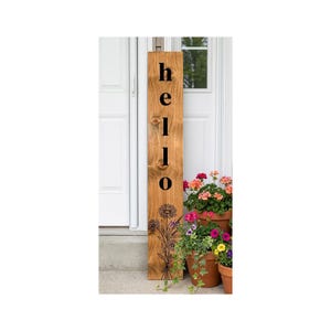 May include: A tall, rectangular wooden sign with a light brown wood grain finish. The word hello is printed vertically in black sans-serif letters. Below the text is a black line art illustration of a cluster of daisies and wildflowers.