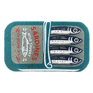 Puede incluir: Felpudo rectangular con borde turquesa y diseño de lata de sardinas. La alfombra presenta cuatro sardinas, una ilustración de pez blanco y las palabras "SARDINES" y "WILD CAUGHT" en rojo. También está presente el texto "IN OLIVE OIL".