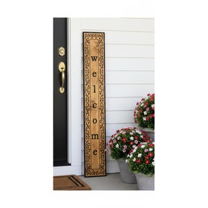 May include: A tall, rectangular wooden sign with a light brown finish and a dark border featuring a black vine pattern. The word "welcome" is printed vertically in black, lowercase, serif lettering down the centre of the wooden board.
