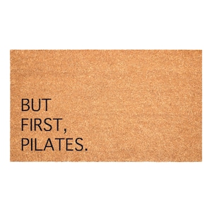 May include: A rectangular doormat with a light brown, textured surface. The black text reads "BUT FIRST, PILATES." This mat is designed for use at a doorway or entrance, and is made of a durable material.