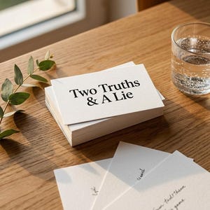 Könnte beinhalten: Ein Stapel weißer Karten mit dem Text "Two Truths & A Lie" liegt auf einem Holztisch, neben einem Glas Wasser und einem Zweig Grün. Mehrere einzelne Karten sind in der Nähe verstreut, was auf ein Spiel hindeutet.