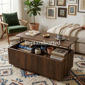 May include: A rectangular dark wood coffee table with a lift-top mechanism, featuring vertical slat detailing on the exterior. The open storage compartment contains folded textiles, ceramic vessels, and books titled Japanese Ceramics and Hygge Living.