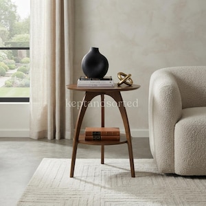 May include: A round wooden side table with three curved legs and a lower shelf. On the top surface sits a matte black vase, a stack of three books, and a gold-toned metal knot sculpture. Two brown books are placed on the lower shelf.
