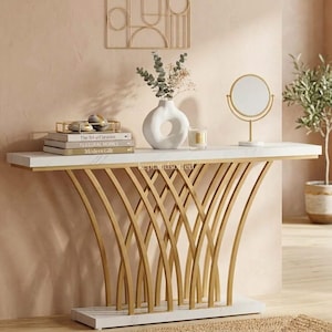 May include: A console table with a white faux marble top and base, featuring a gold-toned metal frame with curved, intersecting supports. On top are three stacked books titled The Art of Curation, Textural Homes, and Modern Gilt, a white vase, and a mirror.