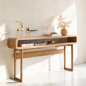 May include: A light wood console table with a rounded rectangular frame and an open shelf. The table features two gold deer figurines, a tan vase with dried flowers, and books on the surface. The text "keptandsorted" is visible on the shelf.