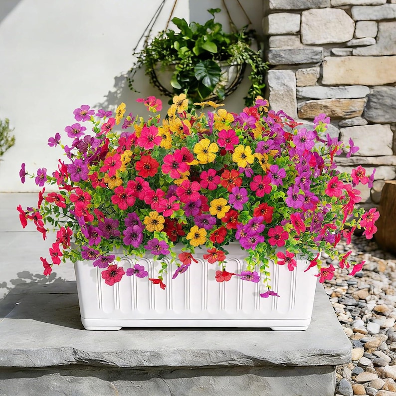 May include: A rectangular white planter box overflowing with vibrant, colorful flowers. The blooms are a mix of red, pink, yellow, and purple. The planter sits on a stone surface, with a hanging basket in the background.