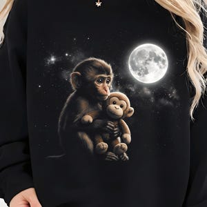 May include: Black sweatshirt featuring a detailed illustration of two monkeys under a full moon and starry night sky. One monkey is holding a stuffed animal monkey. The design is centered on the front of the shirt.