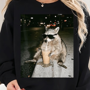 May include: Black sweatshirt featuring a raccoon wearing sunglasses and drinking an iced coffee. The raccoon is sitting on a concrete ledge with a city street background. The image has a nighttime setting with blurred lights.