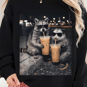 May include: Black sweatshirt featuring a graphic of two raccoons wearing sunglasses, holding iced coffee drinks with straws. The raccoons are depicted in a nighttime urban setting. The sweatshirt is a solid black color.