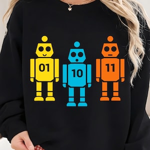 Op de afbeelding: Zwart sweatshirt met drie kleurrijke robotafbeeldingen. De robots zijn geel, blauw en oranje, elk met een ander nummer: 01, 10 en 11. De robots hebben een eenvoudig, geometrisch ontwerp.