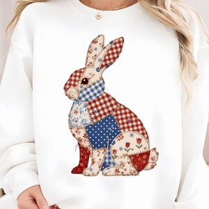 Pode incluir: Camisola branca com um design de coelho em patchwork. O coelho é composto por vários padrões de tecido, incluindo xadrez vermelho, bolinhas azuis e estampas florais. A camisola tem gola redonda e mangas compridas.