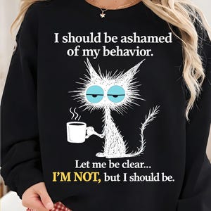 Può includere: Felpa nera con un gatto dei cartoni animati che tiene una tazza di caffè. Il testo recita: "Dovrei vergognarmi del mio comportamento. Sia chiaro... NON LO SONO, ma dovrei."