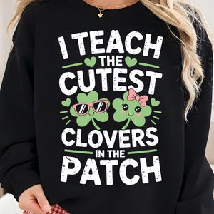 Może przedstawiać: Czarna bluza z białym napisem "I TEACH THE CUTEST CLOVERS IN THE PATCH". Projekt zawiera dwie koniczyny z kreskówek, jedną w okularach przeciwsłonecznych, a drugą z różową kokardą. Bluza ma motyw Dnia Świętego Patryka.