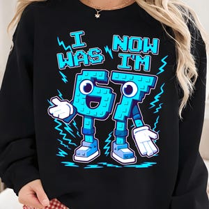 Op de afbeelding: Zwart sweatshirt met de tekst "I WAS 67 NOW I'M 67" in lichtblauw. De cijfers zijn ontworpen als cartoonfiguren met armen, benen en gezichten. Het ontwerp bevat bliksemaccenten.
