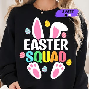 Puede incluir: Sudadera negra con la frase "EASTER SQUAD" en blanco y colores arcoíris. El diseño incluye orejas de conejo, huevos de Pascua y patas de conejo. Un diseño festivo y colorido para la Pascua.