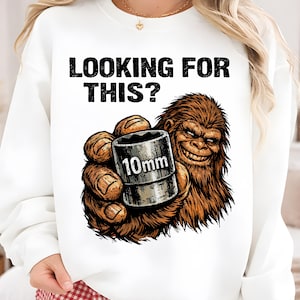 Puede incluir: Sudadera blanca con el texto "LOOKING FOR THIS?" sobre una ilustración de dibujos animados de un Bigfoot sosteniendo un dado de 10 mm. El Bigfoot es marrón y tiene una sonrisa traviesa.