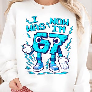 May include: White sweatshirt with a cartoon graphic. The graphic features the text "I WAS 67 NOW I'M 67" in blue pixelated letters. The numbers are personified with arms, legs, and faces. Blue lightning bolts and white gloves complete the design.