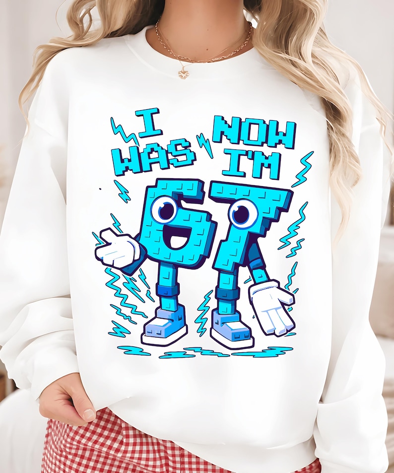 May include: White sweatshirt with a cartoon graphic. The graphic features the text "I WAS 67 NOW I'M 67" in blue pixelated letters. The numbers are personified with arms, legs, and faces. Blue lightning bolts and white gloves complete the design.