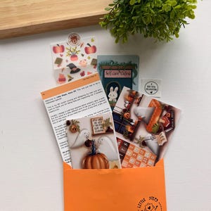 May include: An orange envelope marked "Please DO NOT BEND" and "LITTLE STEPS MAIL CLUB" containing a collection of stationery items including a letter, a calendar, stickers, and postcards featuring ghosts, pumpkins, and a "Self-care Cafe" sign.