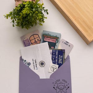 May include: A light purple envelope containing various cards and stickers. The envelope features a cartoon character in an envelope and the text "Little Steps". Included items have text such as "Self-care Cafe", "Today, I choose me", and "June".