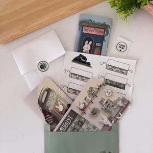 May include: A stationery set featuring a sage green envelope labelled Little Steps Mail Club, a Self-care Cafe card with a white rabbit, a calendar page for August, a blueberry tart illustration, and various cards with prompts like Open When You Feel Sad.