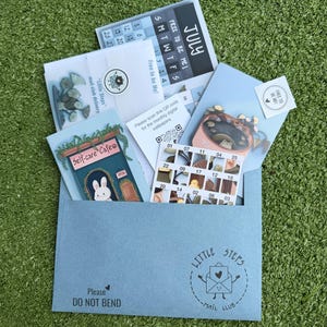 May include: A light blue envelope marked Little Steps Mail Club with a Do Not Bend stamp, containing a July calendar, a Self-care Cafe card featuring a rabbit, a sticker sheet with numbered squares, and a QR code card for digital wallpapers.
