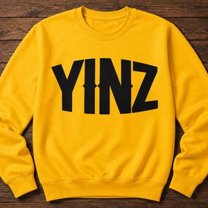 Könnte beinhalten: Ein gelbes Sweatshirt mit dem Wort "YINZ" in großen, schwarzen Buchstaben auf der Brust. Das Sweatshirt hat einen Rundhalsausschnitt und lange Ärmel. Der Hintergrund ist eine dunkelbraune Holzoberfläche.