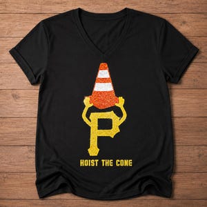 Peut inclure: T-shirt noir à col en V avec un motif de cône de signalisation sur un "P" jaune, avec le texte "HOIST THE CONE" en dessous. Le cône est orange avec des rayures blanches. Le motif a une texture pailletée.