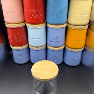 May include: A collection of twelve ceramic jars in various colors including pink, red, blue, yellow, and purple, each topped with a light wood lid. The jars feature embossed text or symbols such as la fermière, Bisou Bisou, Sagittarius, Gemini, and Pisces.