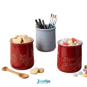 May include: Three ceramic jars, one red with "Be Mine" text and heart-shaped cookies, one gray with art supplies, and one red with "Bisou Bisou" text and heart candies. A wooden spoon with a heart-shaped end is also present.