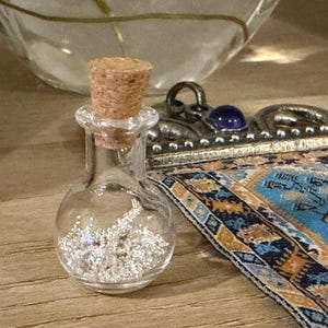 May include: A small, clear glass bottle with a cork stopper, filled with tiny silver-colored metallic particles. Beside it lies a rectangular metal pendant featuring a blue stone and a blue, gold, and black patterned fabric inlay.