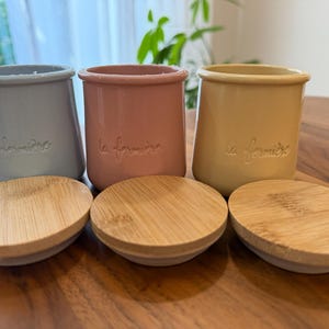 May include: Three ceramic pots in light blue, dusty pink, and mustard yellow, each embossed with the text la fermière. Three round bamboo lids are placed in front of the pots, showing a natural wood grain pattern and a smooth, circular design.