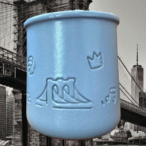 May include: A light blue ceramic mug with embossed designs. The mug features a Brooklyn Bridge illustration, a crown, musical notes, and other symbols. The mug has a rounded base and a slightly flared rim. The background is a black and white image of the Brooklyn Bridge and New York City skyline.