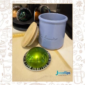 May include: A light blue ceramic jar with the text "la fermière" in cursive, a wooden lid, and a green coffee pod. Several other coffee pods are visible in the background. The jar is likely for storing coffee pods.