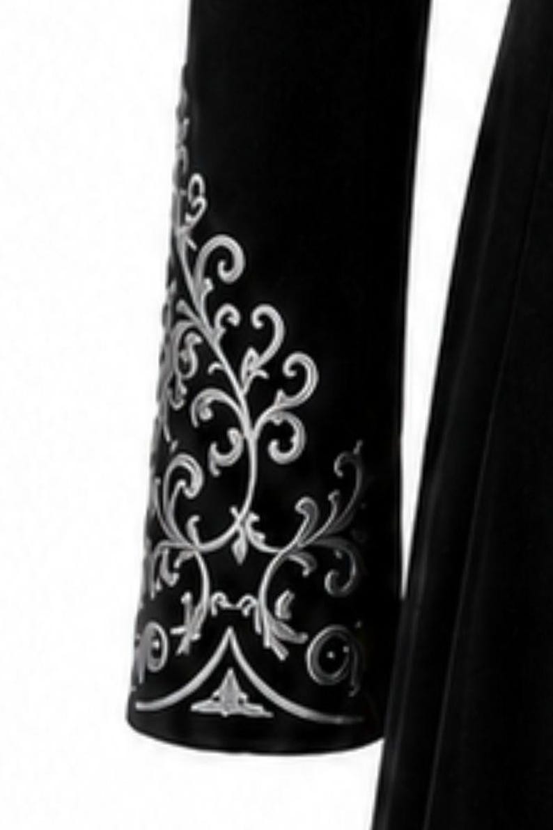 May include: Close-up of a black garment sleeve with detailed silver floral embroidery. The design features swirling vines and leaves, creating a decorative pattern. The sleeve is made of a dark, possibly velvet, material.