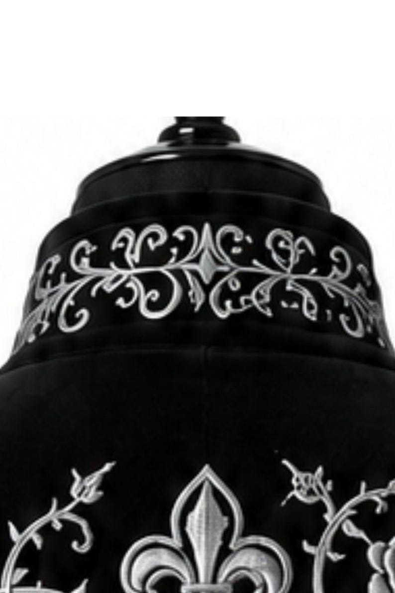 May include: A close-up of a black garment with silver embroidery. The design includes scrolling floral patterns and a fleur-de-lis motif, giving a gothic or regal appearance. The garment seems to be made of a soft, dark fabric.