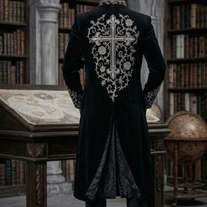 May include: A long black coat with silver cross embroidery on the back. The coat has a high collar and detailed cuffs. The person is in a library with bookshelves and a wooden desk. The coat's design is Gothic-inspired.