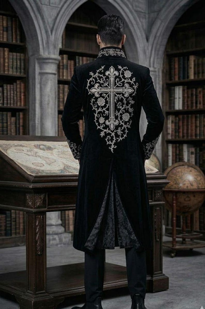 May include: A long black coat with silver cross embroidery on the back. The coat has a high collar and detailed cuffs. The person is in a library with bookshelves and a wooden desk. The coat's design is Gothic-inspired.
