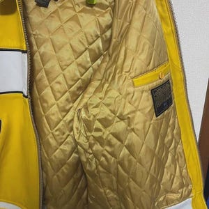 May include: Yellow leather jacket with white stripes and a white hem. The interior is quilted gold fabric. The jacket has a zipper closure and a small pocket. The jacket has the text "BY AVIREX LTD" on the label.