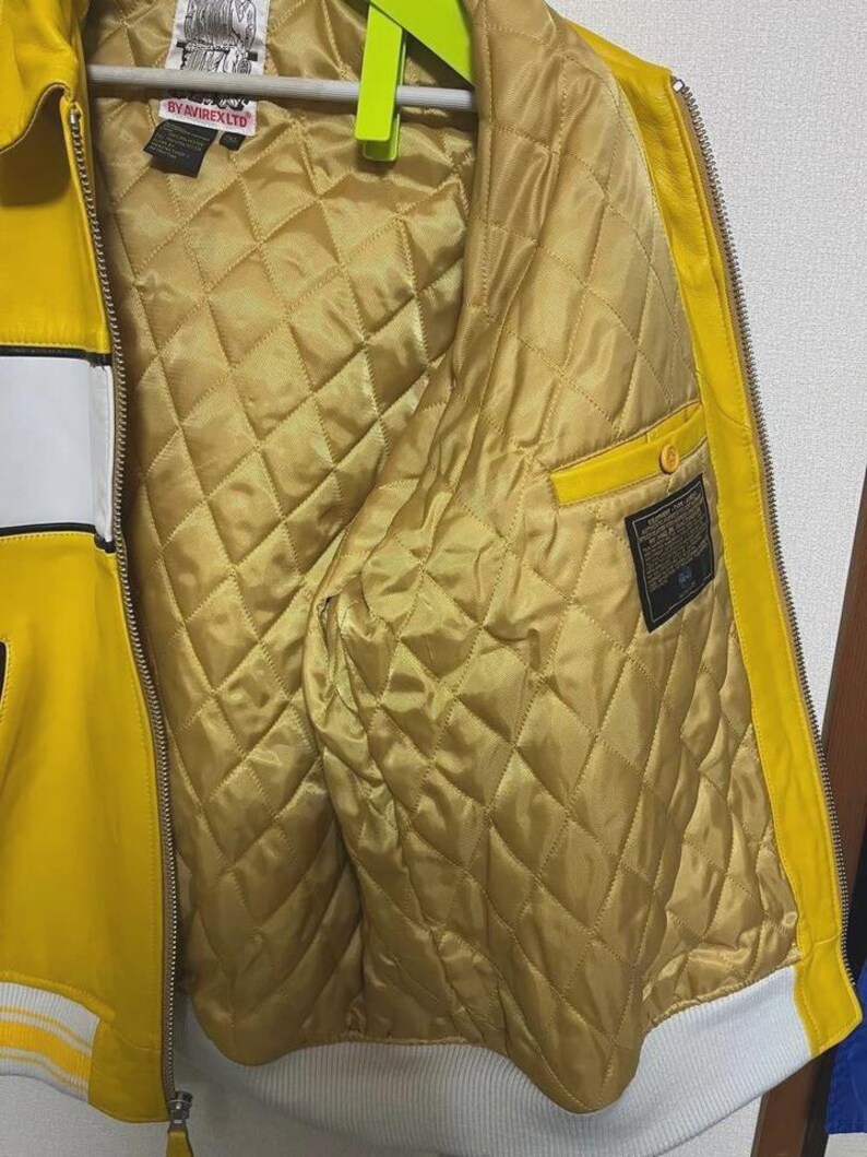May include: Yellow leather jacket with white stripes and a white hem. The interior is quilted gold fabric. The jacket has a zipper closure and a small pocket. The jacket has the text "BY AVIREX LTD" on the label.