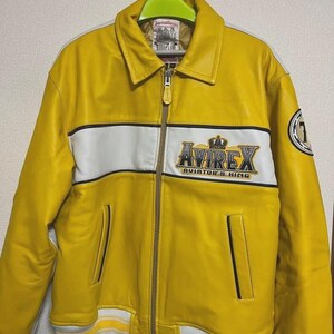 May include: A bright yellow leather jacket with a white stripe across the chest and the text "AVIREX AVIATOR'S KING". The jacket has a zip closure, two front pockets, and a ribbed hem and cuffs. A patch with the number "1" is on the sleeve.