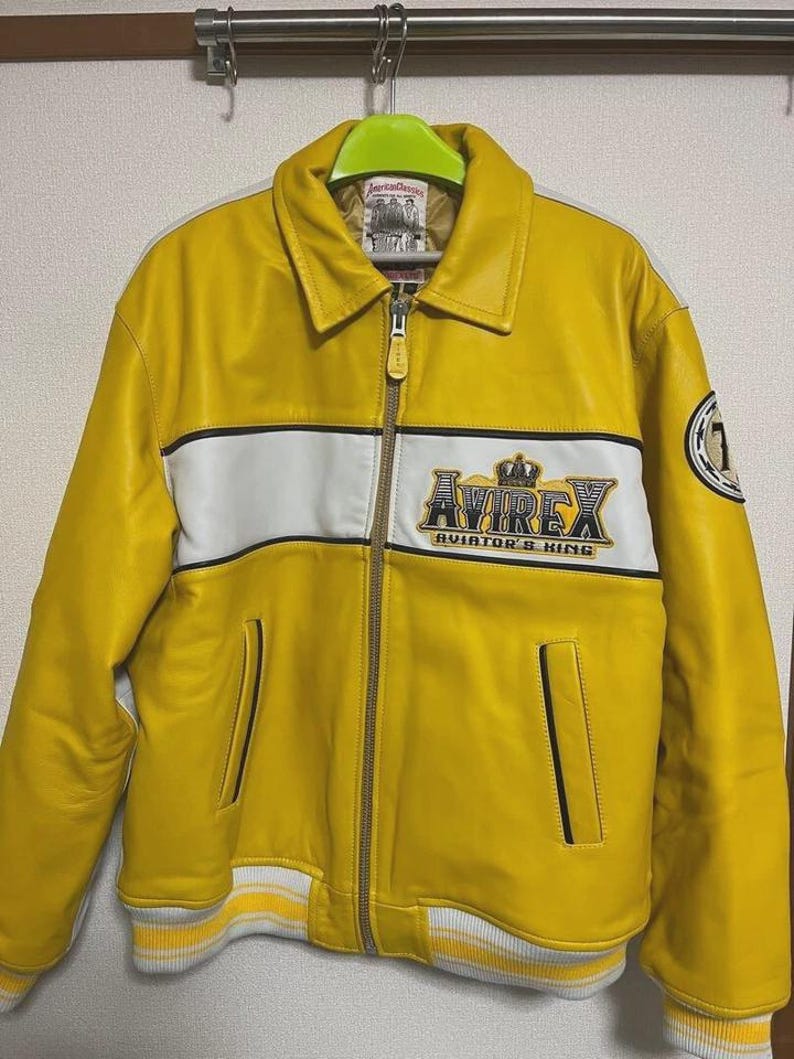 May include: A bright yellow leather jacket with a white stripe across the chest and the text "AVIREX AVIATOR'S KING". The jacket has a zip closure, two front pockets, and a ribbed hem and cuffs. A patch with the number "1" is on the sleeve.