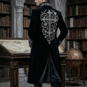 May include: Black velvet coat with silver embroidery of a cross on the back. The coat has a high collar and decorative cuffs. The long coat features a split back. The background shows a library setting with bookshelves and a globe.