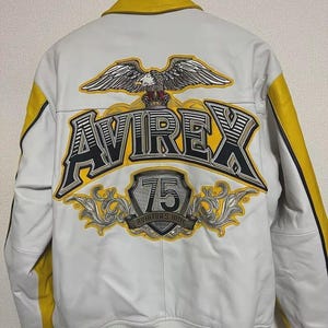 May include: White leather jacket with yellow accents and a yellow collar. The back features a large eagle graphic above the word "AVIREX" in black and yellow, with a "75" emblem below. The jacket has a ribbed yellow and white striped hem.