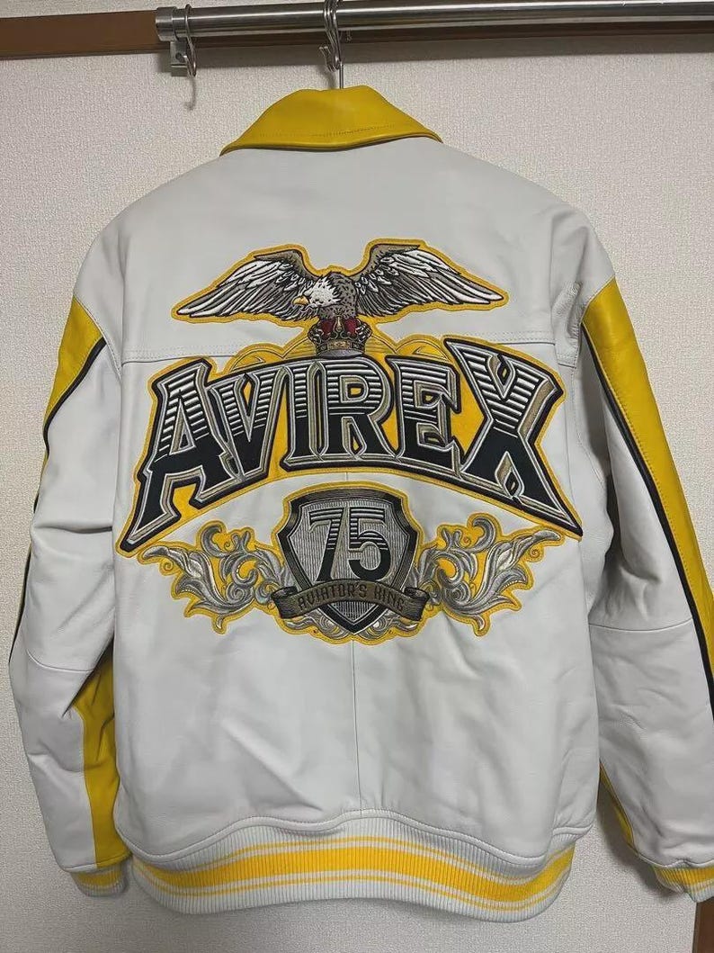 May include: White leather jacket with yellow accents and a yellow collar. The back features a large eagle graphic above the word "AVIREX" in black and yellow, with a "75" emblem below. The jacket has a ribbed yellow and white striped hem.