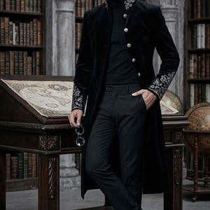May include: A black velvet coat with silver embroidered cuffs and collar. The coat has large silver buttons and is worn over a black turtleneck and black trousers. The setting is a library with bookshelves and a wooden desk.