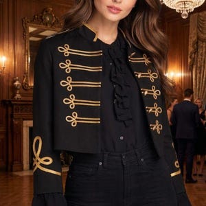 Könnte beinhalten: Schwarze, kurze Jacke mit goldfarbenen Stickereien. Die Jacke hat einen Stehkragen und goldfarbene Verzierungen. Die Ärmel haben eine ausgestellte Manschette. Die Jacke wird mit einer schwarzen Bluse und schwarzen Jeans getragen. Der Hintergrund ist ein Raum mit Kronleuchter.