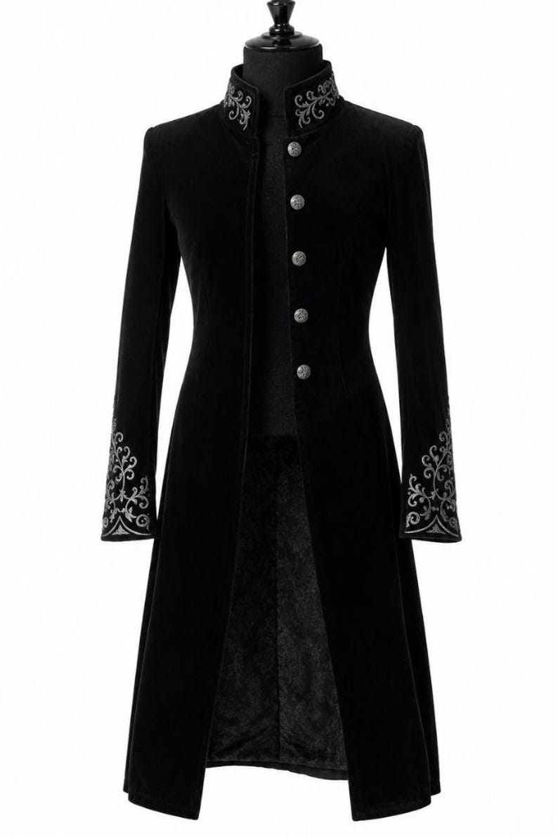 May include: A long, black velvet coat with a high collar and silver embroidered detailing. The coat features silver buttons down the front and ornate embroidery on the cuffs. The coat is displayed on a mannequin.