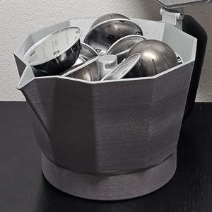 May include: A gray, 3D-printed coffee pod holder shaped like a stovetop espresso maker. The lid is open, revealing several coffee pods inside. The holder sits on a black surface against a white wall.